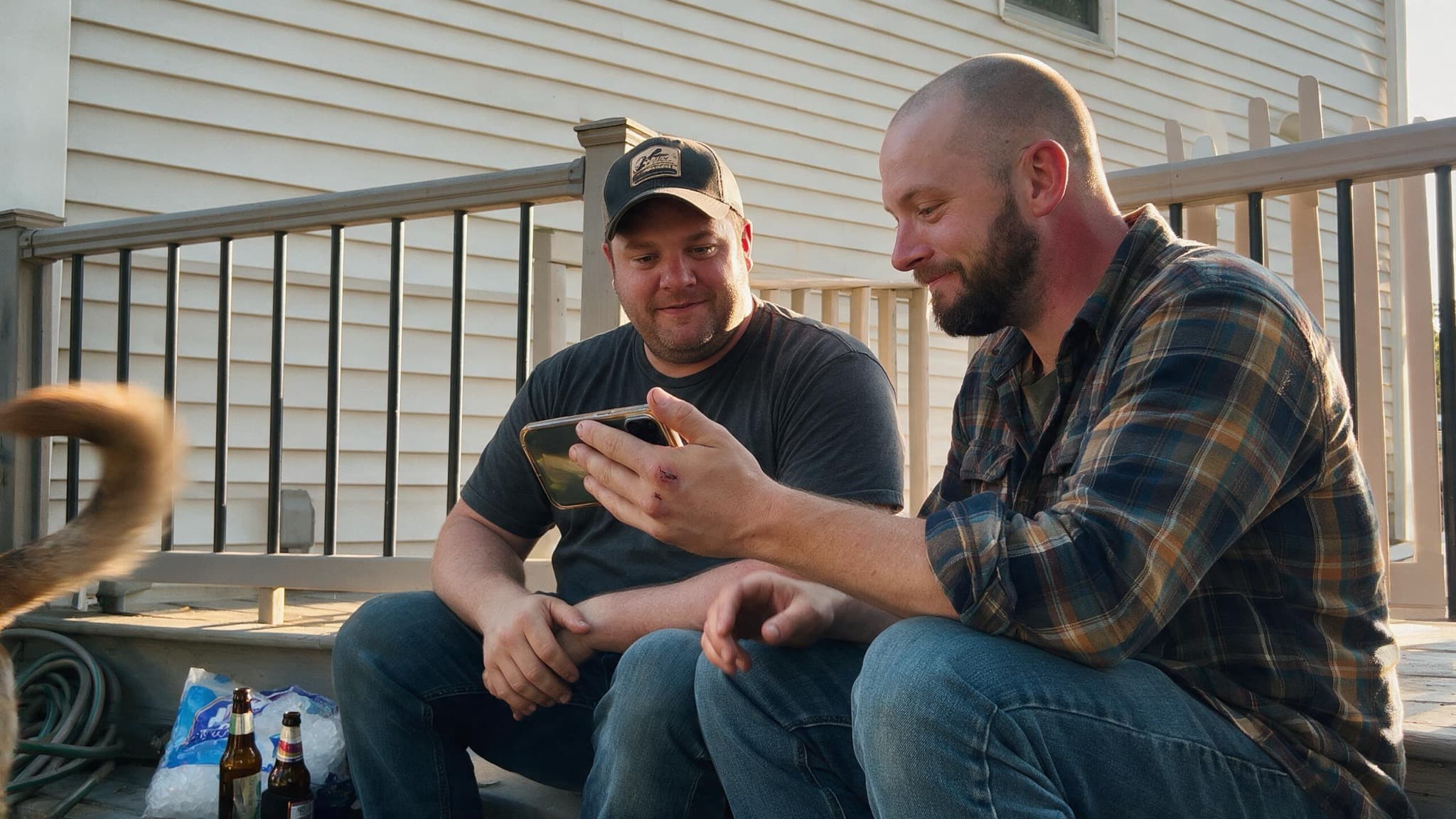 Two brothers in their thirties on a back porch, one showing the other something on a phone
