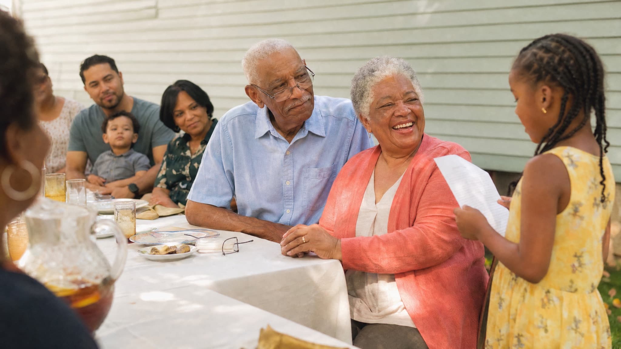 A couple in their seventies at a backyard family lunch, laughing as their granddaughter reads to them from a sheet of paper
