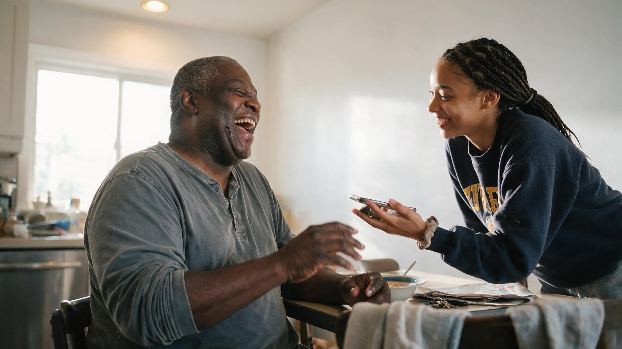 A man in his fifties laughing at a sunlit kitchen table while his daughter leans in to show him something on a phone