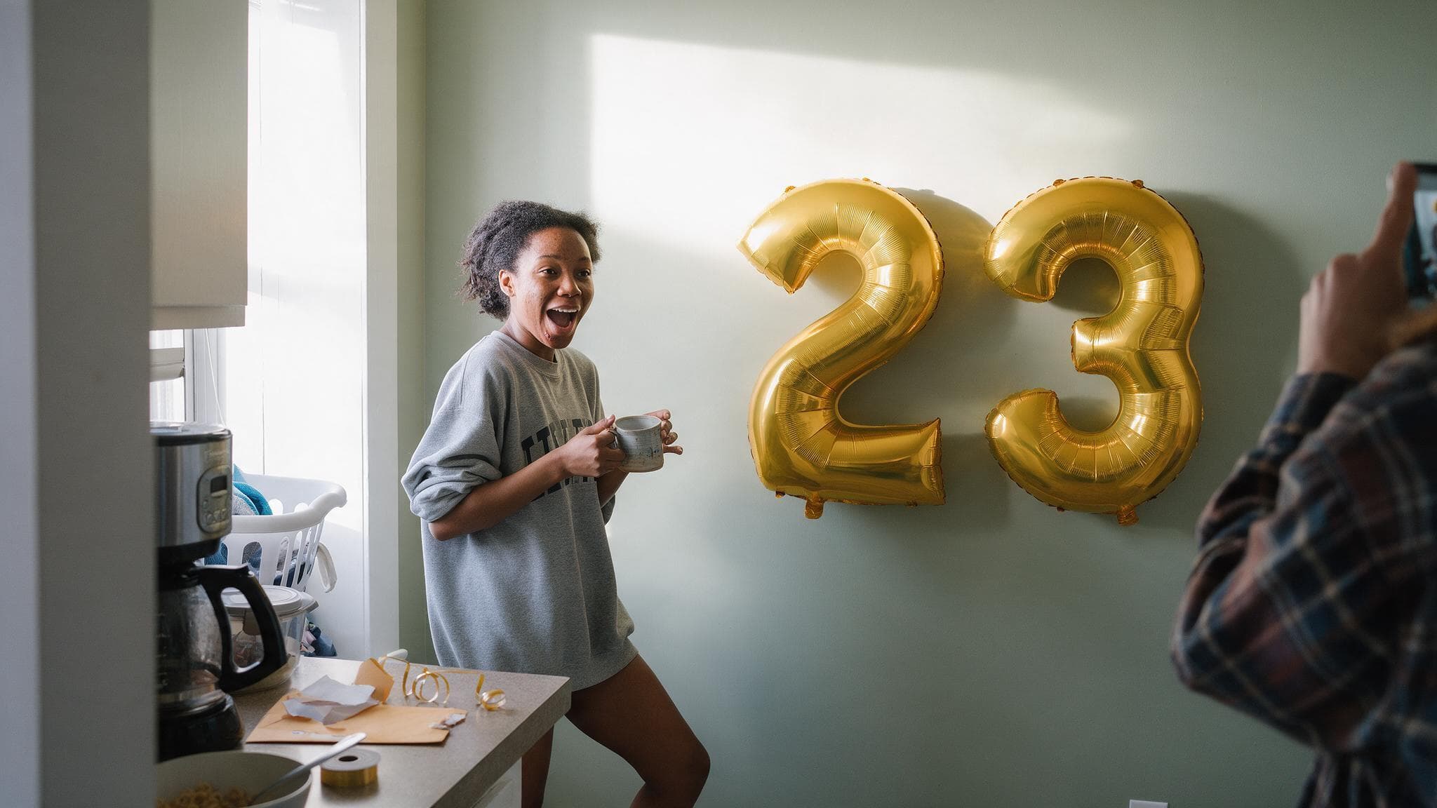 A young woman in a sweatshirt laughing in a bright kitchen at two gold foil number balloons taped to the wall