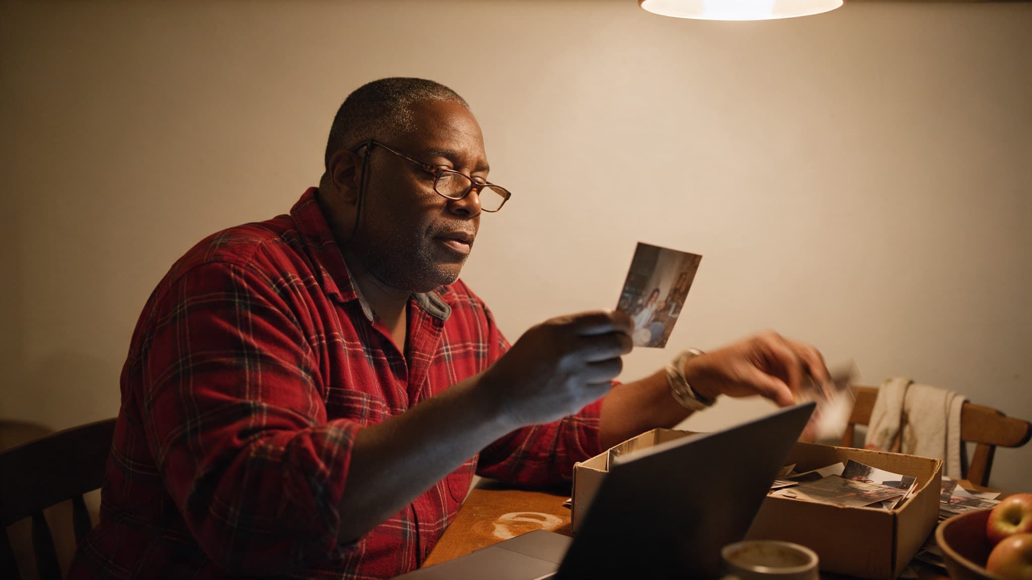 A man at a kitchen table at night holding up an old photograph, a box of loose prints and a laptop in front of him