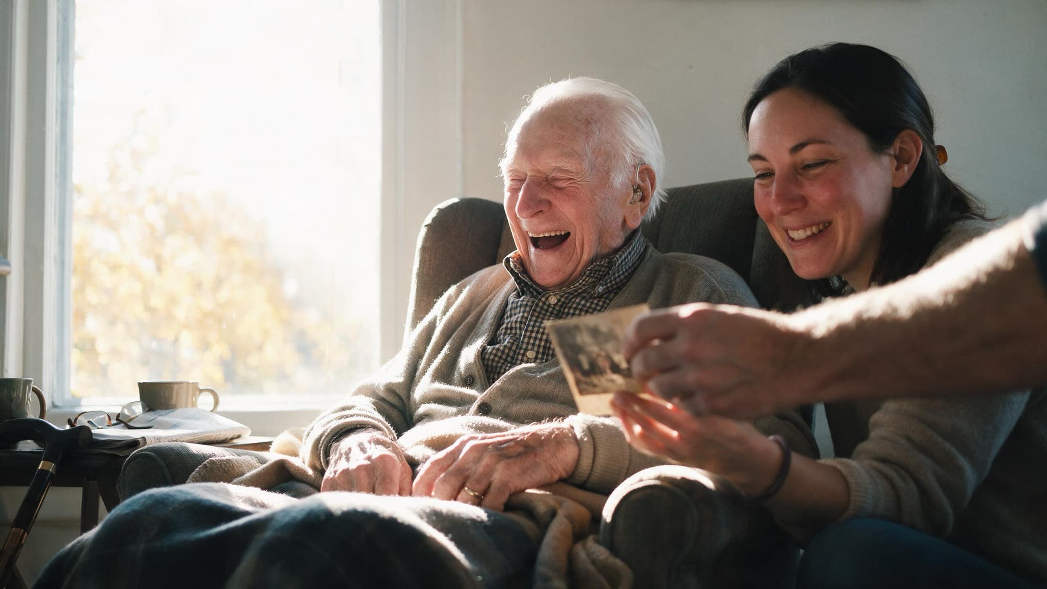 A man in his nineties laughing in an armchair by a sunlit window while his granddaughter leans in beside him with an old photograph