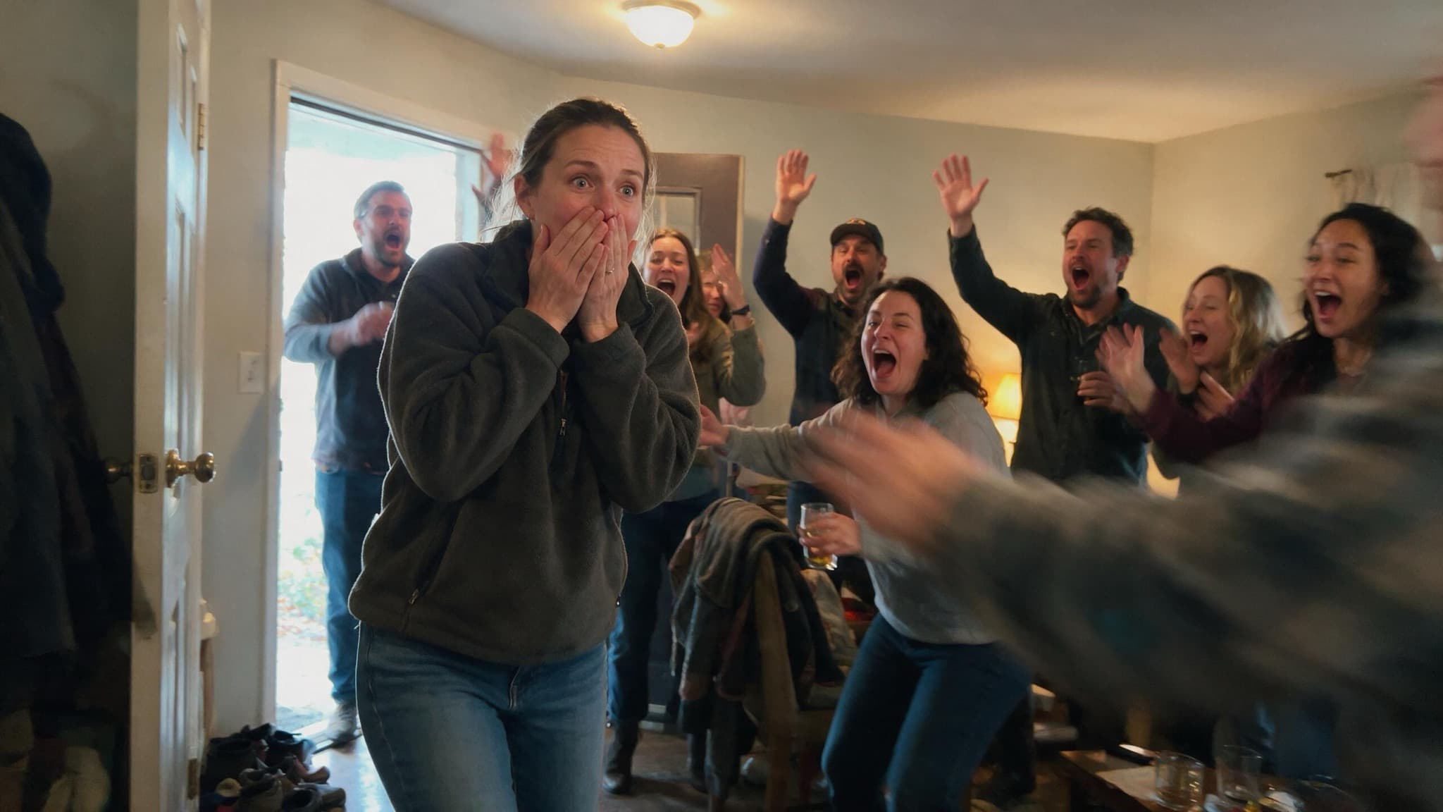 A woman stopping in a doorway with both hands over her mouth as a room of friends turns toward her